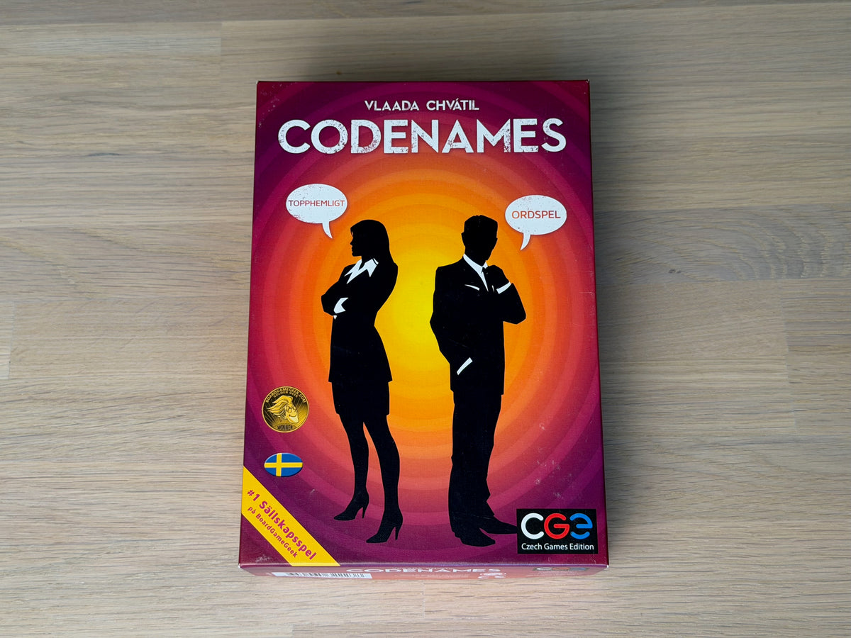 Codenames svenska ask liggande på ett ekgolv, sedd uppifrån.