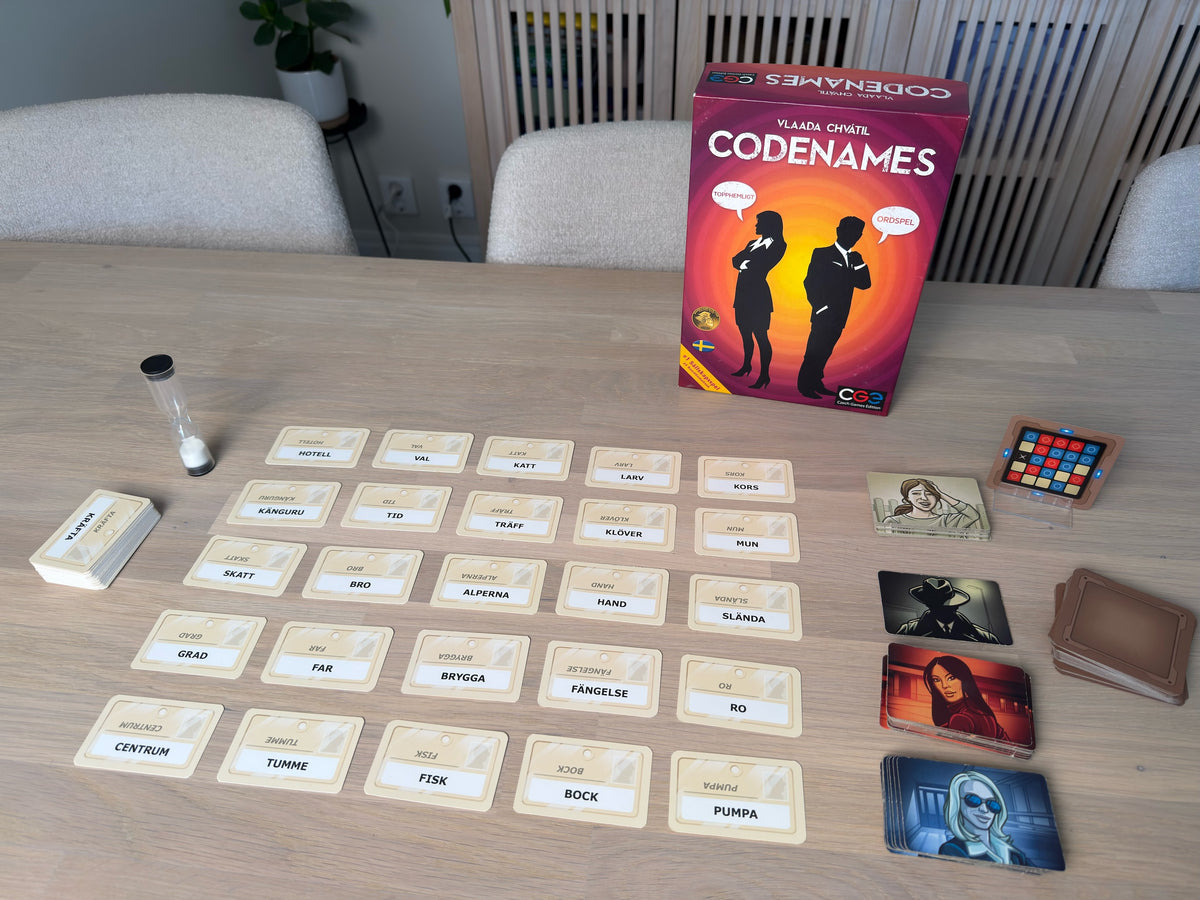 Codenames uppdukat: rutnät med ordkort, timglas, nyckelkort och asken bakom.
