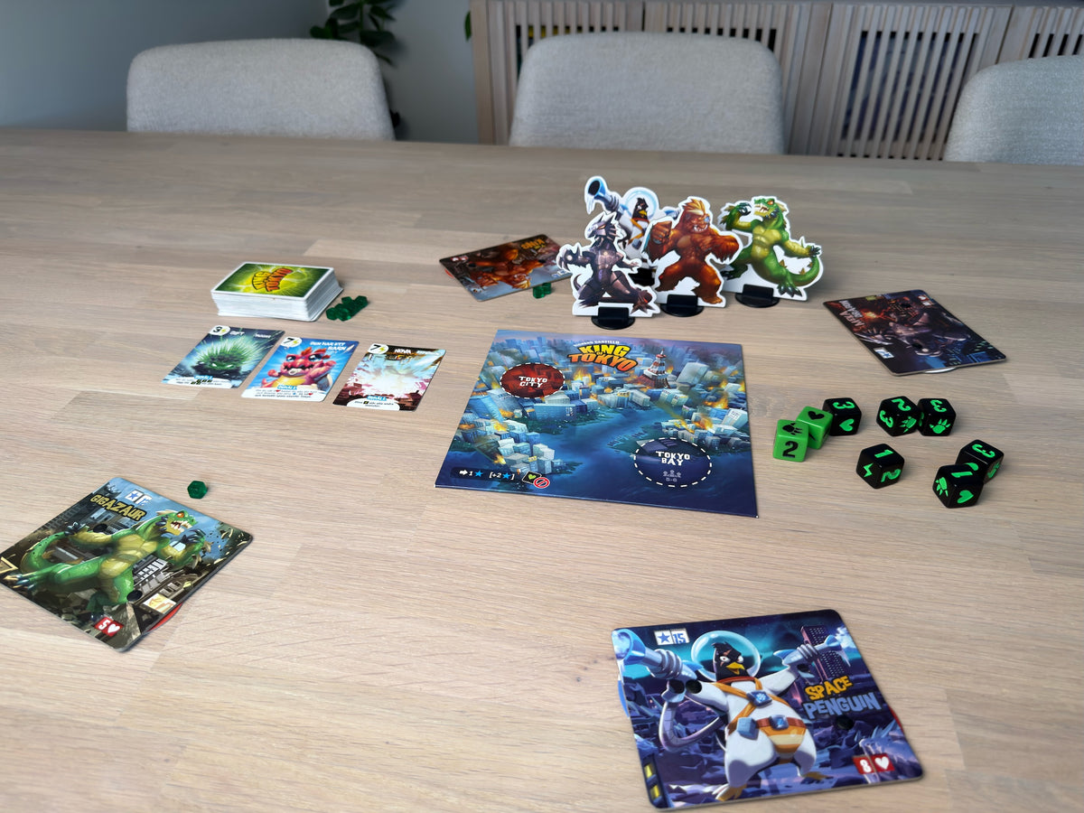 King of Tokyo uppdukat på ett matbord: spelplanen i mitten, monsterfigurerna bakom, tärningar och svenska kort runt omkring.