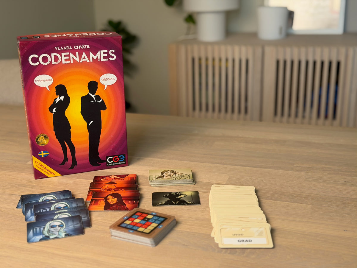 Codenames svenska ask stående på ett träbord med spelet uppdukat framför.